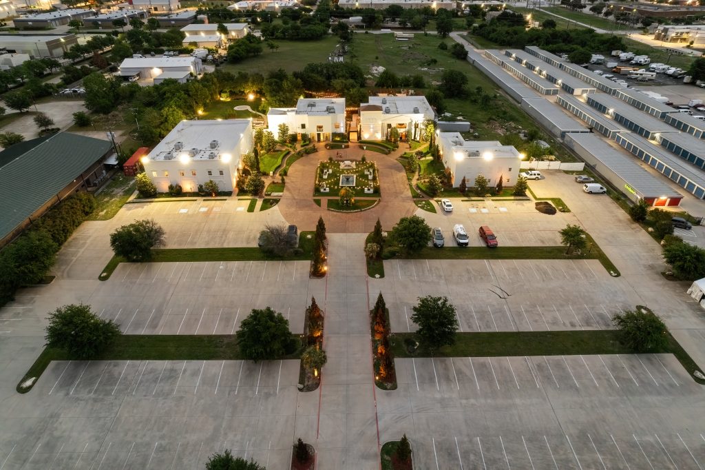 Aerial view of a grand event venue with illuminated pathways, well-manicured gardens, and ample parking space.
